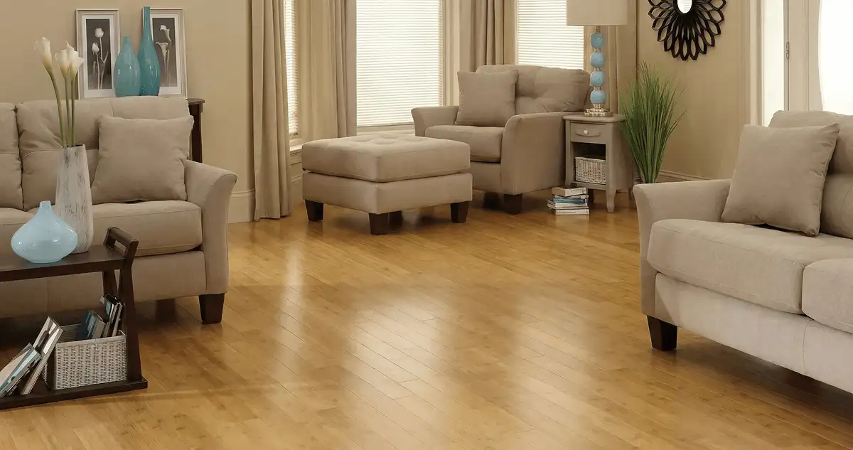 A living room with professionally installed bamboo flooring, beige sofas, a matching ottoman, a side table with a blue lamp, vases, framed pictures, and large windows with blinds and curtains in Stamford, CT